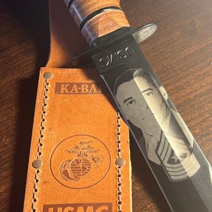 Op de afbeelding: Een bruine leren schede met de woorden "KA-BAR" en "USMC" erop gegraveerd. Een zwart mes met een bruin handvat en een gegraveerd gezicht van een persoon zit in de schede.