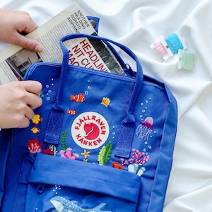 May include: A blue Fjällräven Kånken backpack with a white whale and other sea creatures embroidered on the front. The backpack has a red Fjällräven logo and the text "Fjällräven Kånken" is embroidered on the front.