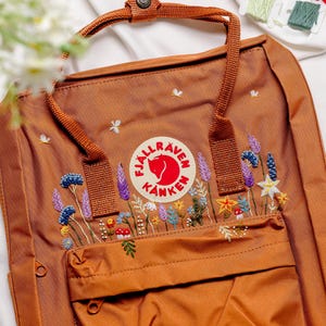 May include: A brown Fjällräven Kånken backpack with a red logo and embroidered flowers in shades of purple, white, and yellow.