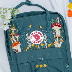 May include: Teal backpack with embroidered mushrooms, yellow flowers, and bees. The backpack features the brand logo and the name "Jenny" embroidered on the front. The design includes red, orange, and brown mushroom details.