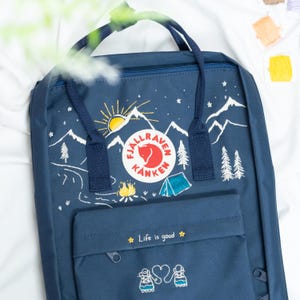 May include: Navy blue backpack with a mountain and sun graphic. The backpack features a circular logo and the text "Life is good". The design includes a tent, campfire, and trees.
