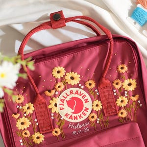 May include: A burgundy backpack with embroidered yellow sunflowers and white accents. The backpack features a circular logo and the name "Halena" with the date "10.11.25" embroidered below. The bag has red straps and a top handle.