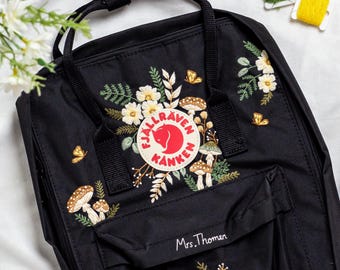 May include: Black backpack with embroidered floral and mushroom design. The backpack features a red and white logo patch and the name "Mrs. Thomen" embroidered on the front pocket. The embroidery includes white flowers, green leaves, and brown mushrooms.