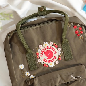 Puede incluir: Mochila verde oliva con bordados florales. La mochila presenta un emblema circular con un logotipo rojo y el texto "Fjallraven Kanken". Tiene un asa superior y correas para los hombros. El nombre "Emma" está bordado en el bolsillo delantero.
