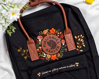 May include: Black backpack with brown leather handles and accents. The front features an embroidered design of autumn leaves, mushrooms, and the text "Fjallraven Kanken." Below, the text "Leaves are falling autumn is calling" is embroidered.