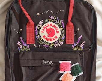 Può includere: Uno zaino nero con manici rossi e dettagli floreali ricamati. Lo zaino presenta un logo circolare e il nome "James" ricamato sul davanti. Tre rocchetti di filo rosa, verde e arancione sono posizionati davanti.