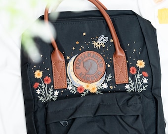 Può includere: Zaino nero con manici e dettagli in pelle marrone. Lo zaino presenta un ricamo di un'ape, fiori e una luna con il logo del marchio. La parte inferiore dello zaino presenta fasi lunari ricamate.