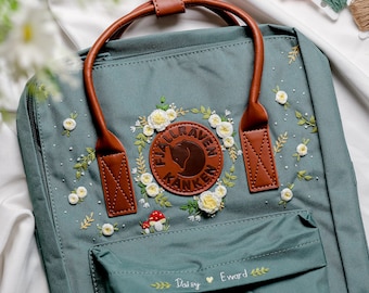 May include: Teal backpack with brown leather handles and accents, featuring embroidered white flowers and green leaves. A circular leather patch displays a brown moose. The text "Daisy Eward" is embroidered on the front.