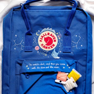 Puede incluir: Una mochila Fjällräven Kånken azul con un diseño bordado blanco de estrellas, una luna y una ballena. La mochila tiene un pequeño bolsillo en la parte delantera con el texto "The world is dark, and then you come with the stars and the moon." bordado en blanco.