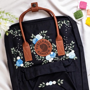 Op de afbeelding: Zwarte rugzak met bruine leren handgreep en riemen, met geborduurde blauwe bloemen en groene bladeren. Een bruine leren patch met de tekst "Fjallraven Kanken" is gecentreerd op de voorkant. De rugzak heeft een voorvak.