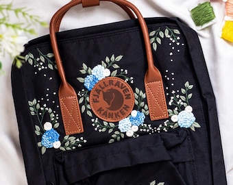 Può includere: Zaino nero con manico e cinghie in pelle marrone, con ricami di fiori blu e foglie verdi. Una toppa in pelle marrone con la scritta "Fjallraven Kanken" è centrata sul davanti. Lo zaino ha una tasca frontale.