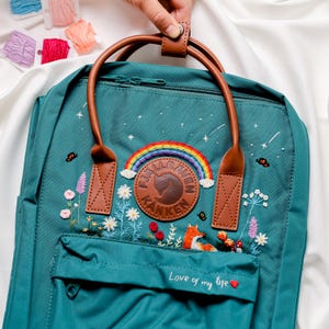 May include: A teal blue Fjällräven Kånken backpack with a rainbow, stars, and flowers embroidered on the front. The backpack has brown leather straps and a brown leather patch with the Fjällräven Kånken logo. The text "Love of my life" is embroidered on the bottom of the backpack.