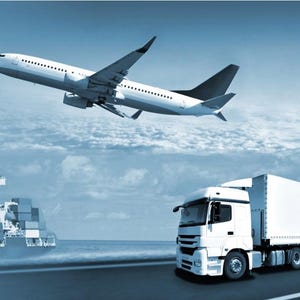 May include: A white airplane flying over a cargo ship and a white semi-trailer truck on a road.