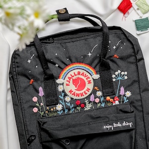May include: Black backpack with embroidered floral and celestial designs. Features a rainbow patch and the text "enjoy little things." The backpack has a top handle, adjustable straps, and a front pocket.