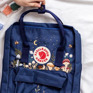 May include: A navy blue backpack with an embroidered forest scene, including mushrooms, flowers, and a snail. The backpack features a top handle, a front pocket, and the Fjallraven Kanken logo. The background has a night sky design with a crescent moon and stars.