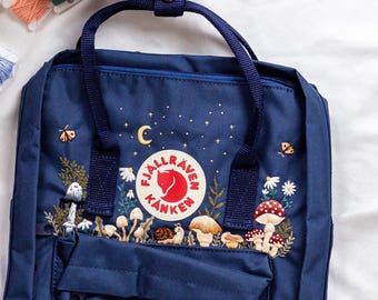 May include: Navy blue backpack with embroidered forest scene featuring mushrooms, flowers, and a snail. The backpack has a top handle, a front pocket, and the Fjallraven Kanken logo. The background has a night sky design with a crescent moon and stars.
