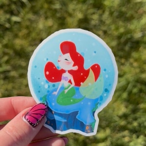 Op de afbeelding: Een gestanste sticker met een cartoon zeemeermin met rood haar en een groene staart in een blauwe sneeuwbol. De zeemeermin zit op een blauwe rotsformatie. De achtergrond is een wazig groen.
