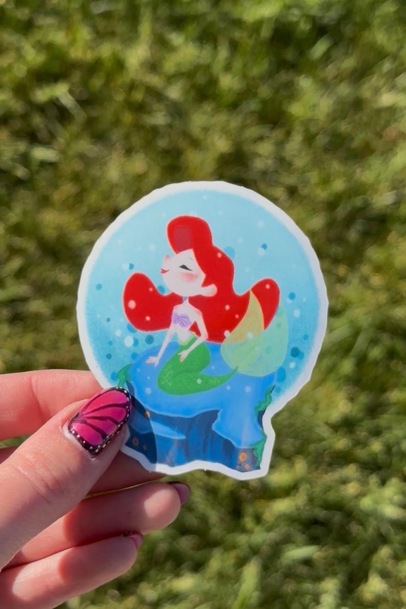 Op de afbeelding: Een gestanste sticker met een cartoon zeemeermin met rood haar en een groene staart in een blauwe sneeuwbol. De zeemeermin zit op een blauwe rotsformatie. De achtergrond is een wazig groen.