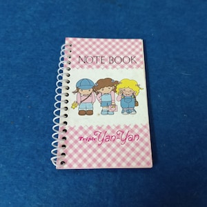 以下が含まれることがあります： ピンクと白のチェック柄のノートで、スパイラル綴じられています。表紙には「NOTE BOOK」という文字と、「Triple Yan-Yan」という文字が書かれており、3人の女の子の漫画が描かれています。