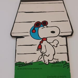 Op de afbeelding: Een gestanst notitieblok in de vorm van een hondenhok met Snoopy die een blauwe vliegeniersmuts met rode bril en een rode sjaal draagt. De afbeelding bevat de tekst Schulz, 1965 United Feature Syndicate Inc en Contents 40 Sheets op een groene basis.