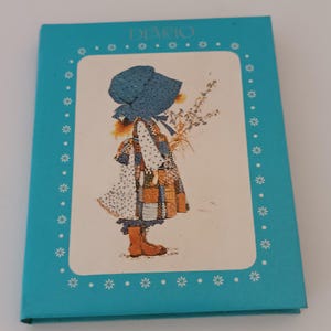 Può includere: Un diario rettangolare color turchese con un pannello centrale bianco che presenta un'illustrazione stampata di un bambino con una cuffietta blu e un vestito patchwork, che tiene in mano un mazzo di fiori. La parola DIARIO è stampata in alto.