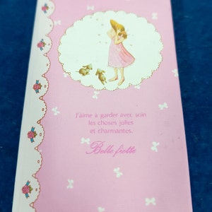 Puede incluir: Una tarjeta rosa y blanca con un diseño floral y lazos. La tarjeta presenta una ilustración de una niña con un vestido rosa con dos perros pequeños. El texto "J'aime à garder avec soin les choses jolies et charmantes. Belle fiette" está impreso en la tarjeta.
