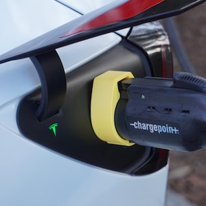 May include: Close-up of a white electric vehicle charging port with a black charging cable. The port has a yellow cover and the text "-chargepoint+" is visible on the charging cable. A green Tesla logo is on the vehicle.
