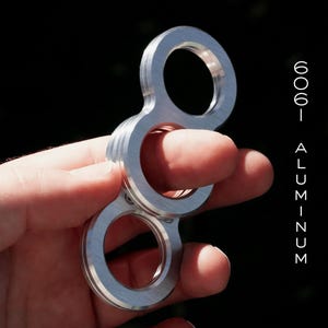 May include: A silver-colored, figure-eight shaped fidget toy made of 6061 aluminum. The toy has two circular openings and is held in a person's hand against a dark background. The text "6061 ALUMINUM" is vertically aligned on the right side.