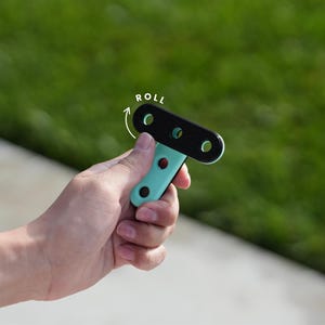 May include: A black and teal T-shaped tool with three circular holes and the word "ROLL" in white. The tool is held in a hand against a blurred green background.