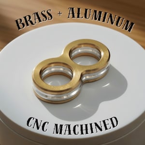 May include: A brass and aluminum CNC machined object in the shape of an infinity symbol. The object has a gold-colored exterior with silver-colored bands. The text "BRASS + ALUMINUM" and "CNC MACHINED" are displayed.