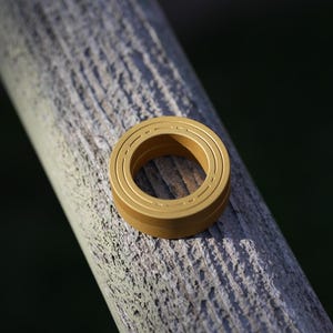 May include: A gold-colored metal ring with three concentric circles, resting on a weathered wooden surface. The ring has a smooth, polished finish and a circular opening in the center. The background is a blurred, dark green.