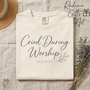 Puede incluir: Camiseta color crema con el texto "Cried During Worship (AGAIN)" en una fuente elegante. La camiseta tiene cuello redondo y mangas cortas. Una ramita de hojas está al lado de la palabra "Worship".