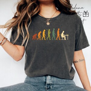 Pode incluir: Uma t-shirt de manga curta cinzento-carv&atilde;o com uma impress&atilde;o gr&aacute;fica da evolu&ccedil;&atilde;o humana, come&ccedil;ando com um primata e progredindo para uma pessoa sentada a uma secret&aacute;ria com um computador. O design utiliza cores laranja, amarelo e verde.