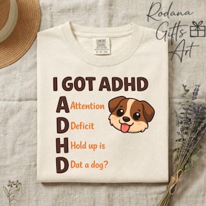 May include: Cream-colored t-shirt with the text "I GOT ADHD" in brown. The acronym is explained with "Attention, Deficit, Hold up is, Dat a dog?" and a cartoon dog illustration. The shirt has a Comfort Colors label.