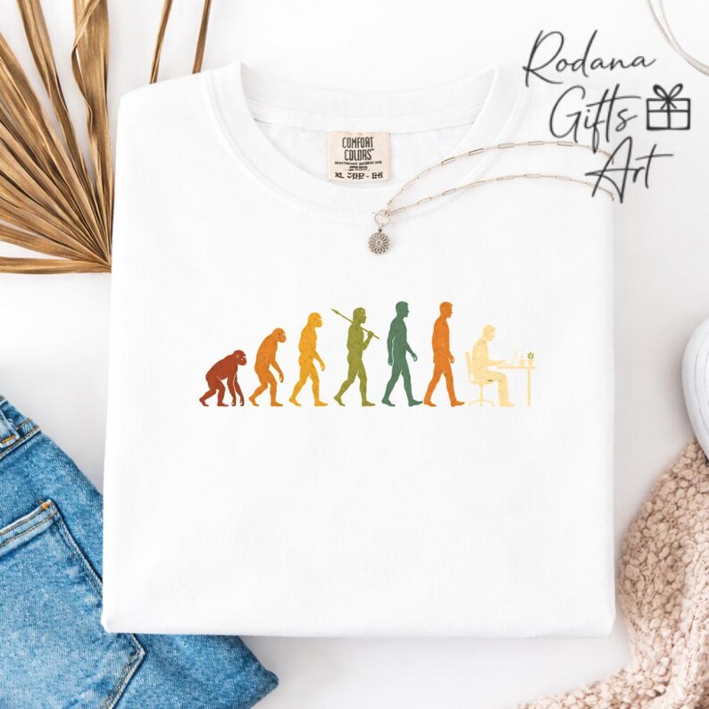 Pode incluir: Uma t-shirt branca com um gr&aacute;fico multicolorido da evolu&ccedil;&atilde;o humana, progredindo de um primata para uma pessoa sentada a uma secret&aacute;ria com um computador. A t-shirt tem um decote redondo e uma etiqueta da marca Comfort Colors no interior.