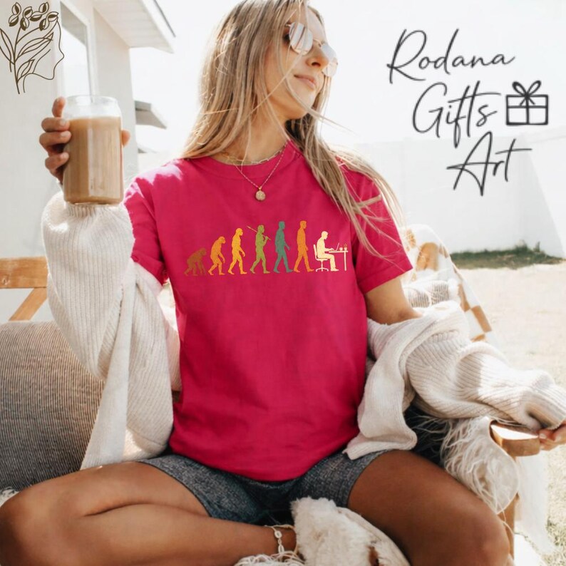 Pode incluir: Uma t-shirt de manga curta cor-de-rosa brilhante com um gr&aacute;fico multicolorido da evolu&ccedil;&atilde;o humana, desde um primata at&eacute; uma pessoa sentada a uma secret&aacute;ria com um computador. O texto Rodana Gifts Art &eacute; vis&iacute;vel no canto superior direito.