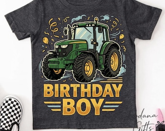 May include: A charcoal grey short-sleeve t-shirt featuring a graphic of a green tractor surrounded by yellow confetti and balloons. Below the tractor, the text "BIRTHDAY BOY" is printed in bold yellow capital letters with horizontal lines.