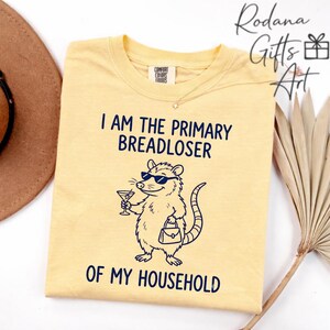 May include: A pale yellow t-shirt featuring a dark blue graphic of an opossum wearing sunglasses, holding a martini glass, and carrying a handbag. The text reads I AM THE PRIMARY BREADLOSER OF MY HOUSEHOLD in dark blue lettering on the front.