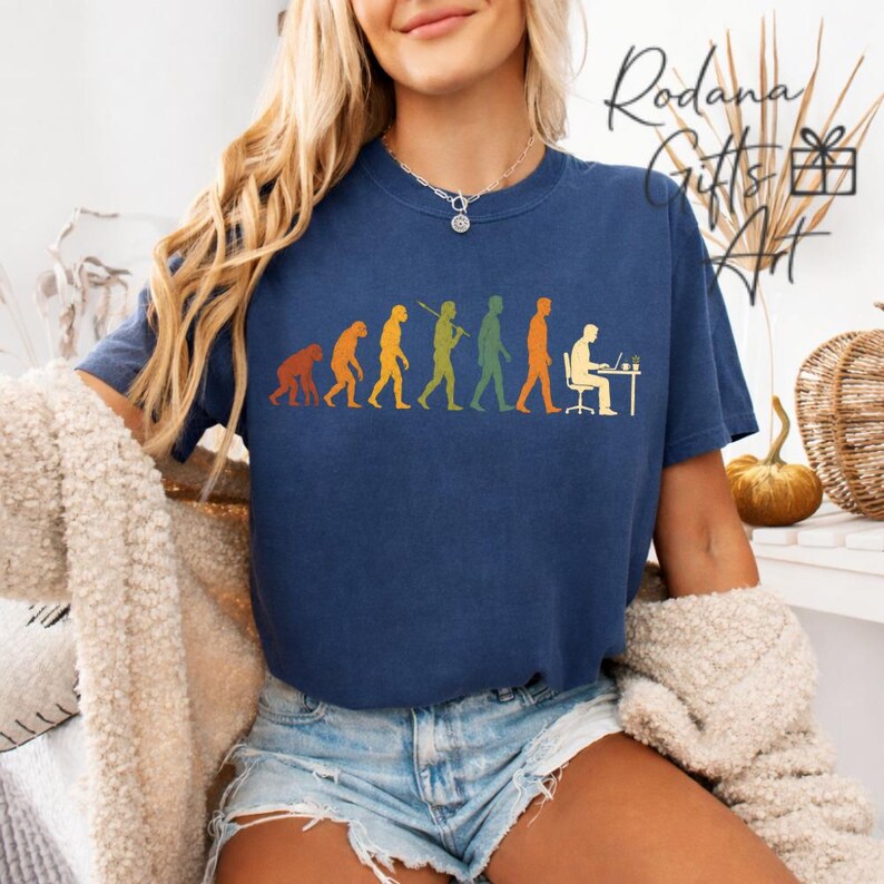 Pode incluir: Uma t-shirt azul-marinho de manga curta com uma estampa gr&aacute;fica da evolu&ccedil;&atilde;o humana, progredindo de um primata para uma pessoa sentada a uma secret&aacute;ria com um computador. O design usa silhuetas laranja, amarelas e verdes na frente.