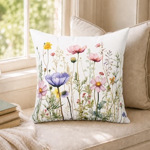May include: A square throw pillow featuring a white background with a colorful watercolor-style print of various wildflowers including purple, pink, and yellow blossoms with green stems and foliage scattered across the front surface of the fabric.