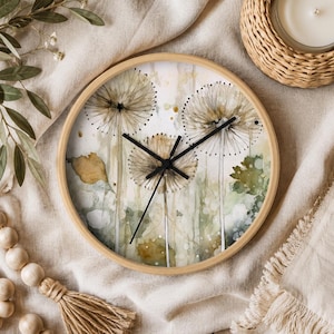 May include: A round wall clock with a light wood frame featuring a watercolor illustration of three dandelion seed heads in shades of brown, green, and white. The clock has three thin black hands centered on the face against a soft botanical background.