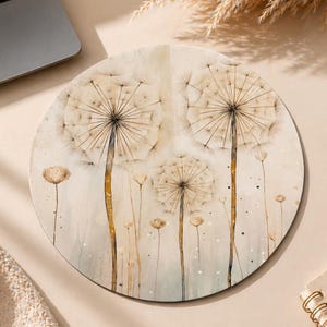 May include: A circular mouse pad featuring a watercolor-style illustration of three large dandelion seed heads with long, thin stems in shades of beige, cream, and soft brown against a light, neutral-toned background with subtle white speckles.