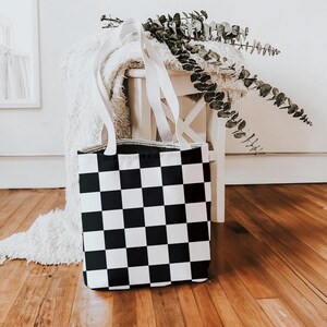 May include: A black and white checkered tote bag with white handles. The bag is sitting on a white fuzzy surface and a white wooden stool.
