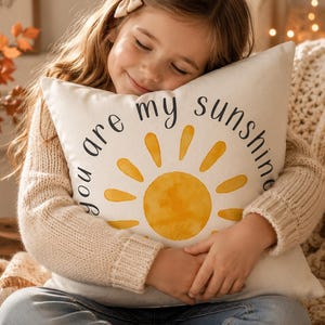 May include: A cream-colored pillow with the words "you are my sunshine" encircling a yellow watercolor sun design. The pillow is being held by a person wearing a cream-colored sweater.