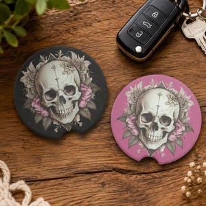 May include: Two circular car coasters featuring a detailed human skull illustration surrounded by pink and grey floral accents. One coaster has a black background, while the other has a pink background. Both display a central skull with a vertical line design.