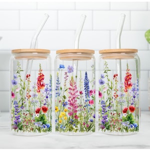 May include: Three clear glass drinking cups with bamboo lids and glass straws. Each cup is decorated with a colorful floral design featuring red, purple, blue, and yellow wildflowers. The cups are displayed on a white surface.