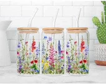 Könnte beinhalten: Drei klare Glastrinkbecher mit Bambusdeckeln und gebogenen Glasstrohhalmen. Jeder Becher ist mit einem farbenfrohen Aquarell-Druck verschiedener Wildblumen in Rosa-, Lila-, Blau-, Rot- und Gelbtönen in einem dichten botanischen Muster versehen.
