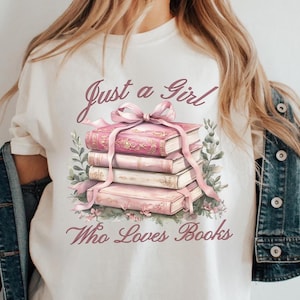 May include: White t-shirt with a graphic of a stack of pink books tied with a pink ribbon, surrounded by greenery and flowers. The text "Just a Girl Who Loves Books" is printed above and below the books.
