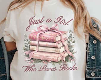 Può includere: T-shirt color crema con un'illustrazione di libri rosa legati con un nastro rosa e dettagli floreali. Il testo "Just a Girl Who Loves Books" è stampato in un carattere corsivo, perfetto per gli amanti della lettura.