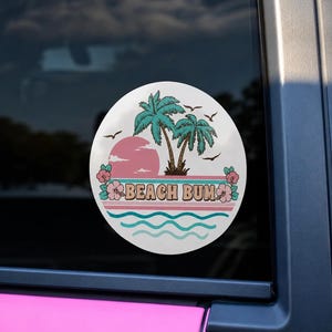 May include: A round sticker with a beach theme. It features a pink sunset, two palm trees, and the words "BEACH BUM" in a retro font. The design includes teal waves, birds, and hibiscus flowers.